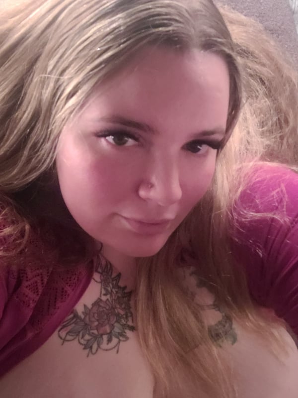 Close-up portrait of a woman with long wavy brown hair and tattoos on her chest, wearing a pink top, smiling softly indoors.