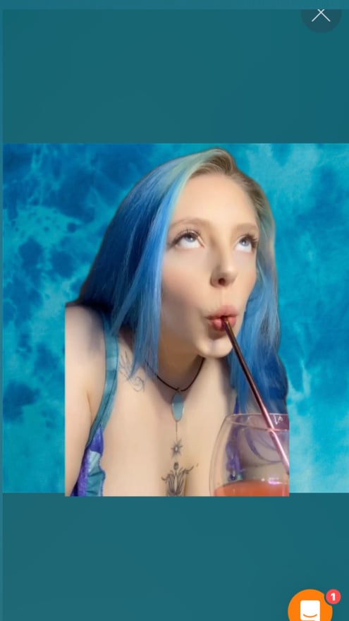 A woman with blue hair sips from a straw while posing playfully against a teal tie-dye background.