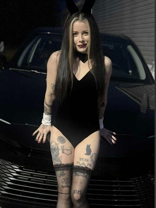 A young woman with long dark hair and bunny ears stands confidently in front of a car at dusk, wearing a black bodysuit with stockings and high heels, displaying tattoos.