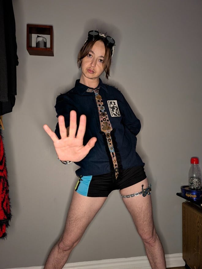 Person standing in a room wearing steampunk goggles, a patterned shirt, tie, and shorts, making a hand gesture in front of a neutral wall.