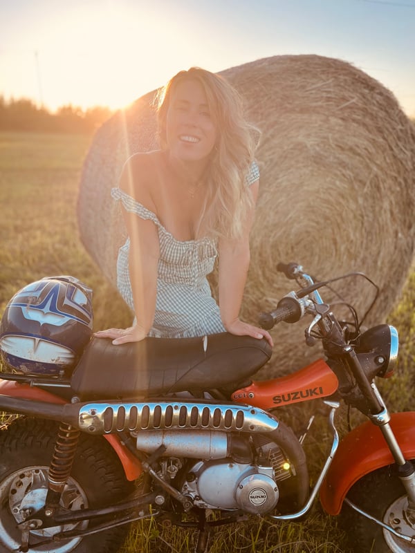 Young woman with long blonde hair smiling happily next to a hay bale and an orange vintage motorcycle at sunset in a field.