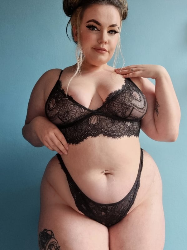 A woman with blonde hair in a bun posing confidently in black lace lingerie against a blue background.