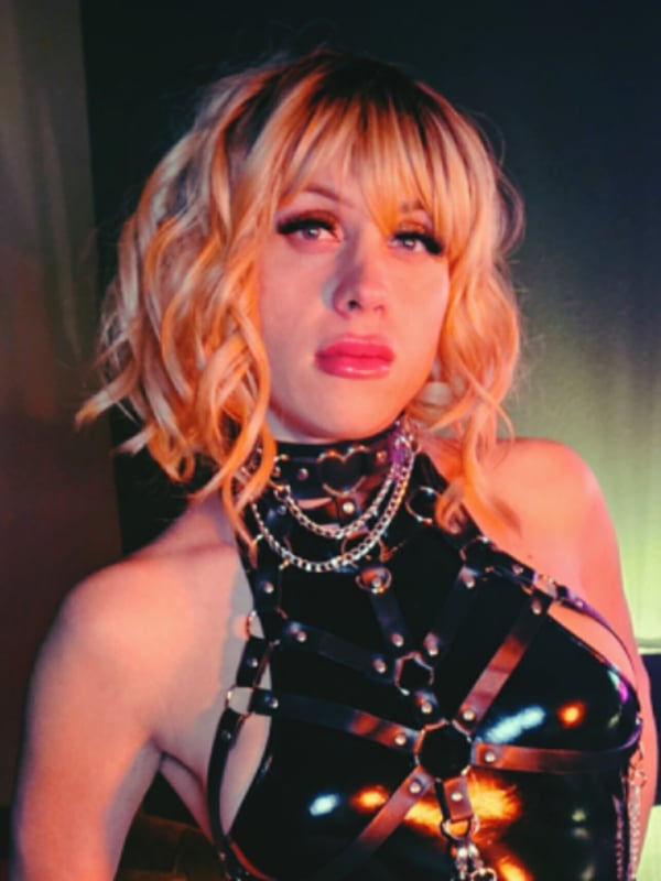 Close-up portrait of a woman with blonde bangs and bold makeup wearing a shiny black harness top against a dark background.