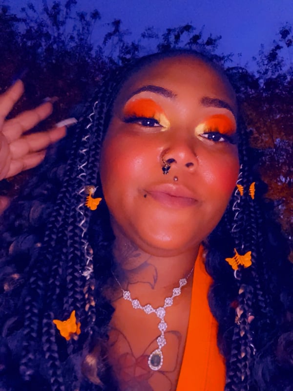 Close-up portrait of a woman with braided hair, orange makeup, and jewelry posing against a twilight outdoor background.