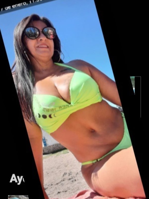 Woman in green bikini posing on the beach with sunglasses, smiling against a blue sky.