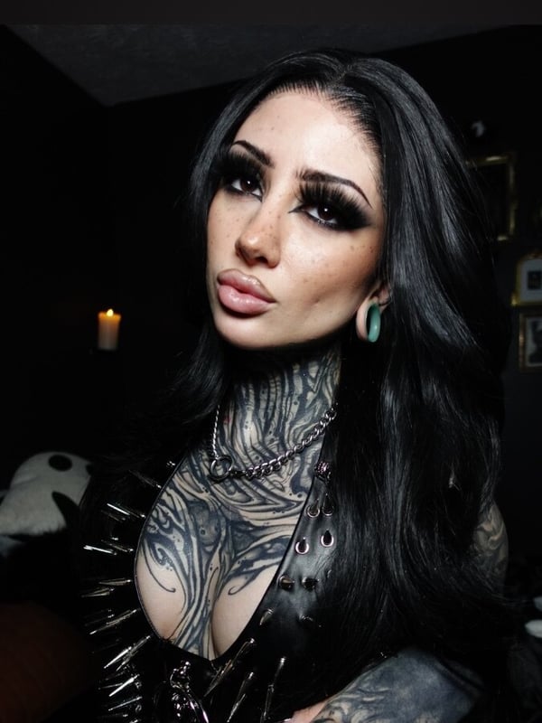 A woman with intricate tattoos, dark eyeliner, and piercings sits in a dimly lit room with gothic decor and a candle.