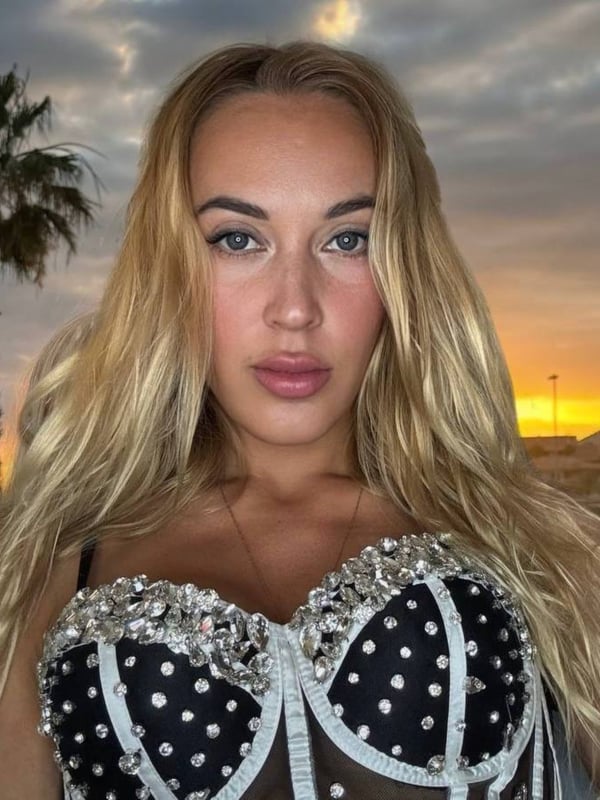 A woman with long blonde hair standing outdoors at sunset near a palm tree, wearing a sparkling black and white strapless top.