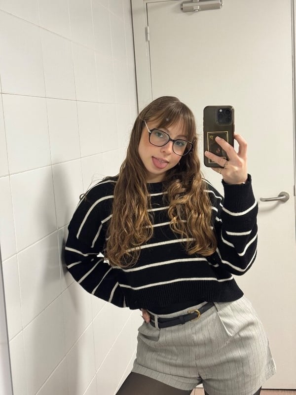 Young woman with long wavy brown hair and glasses sticking out her tongue in a mirror selfie in a bathroom, dressed in a black and white striped sweater and gray skirt with sheer stockings.