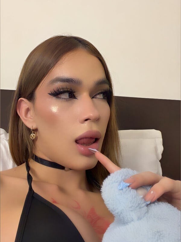 Person with long brown hair, bold makeup, and hoop earrings posing playfully with a blue plush toy indoors.