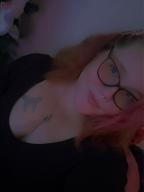 Close-up portrait of a woman with red hair and glasses smiling softly, with holiday decorations and a plush toy in the background.