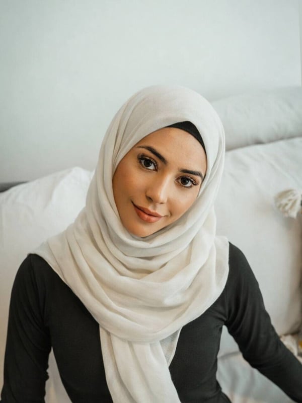 A woman wearing a white hijab and black top, smiling softly against a white background with pillows.