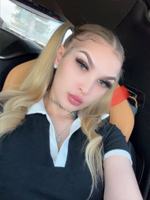 A blonde woman with pigtails sits in a car wearing a collared shirt, smiling at the camera.