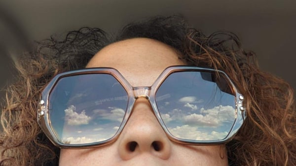 Close-up portrait of a woman wearing large reflective sunglasses with a cloudy sky mirrored in the lenses, curly hair framing her face.