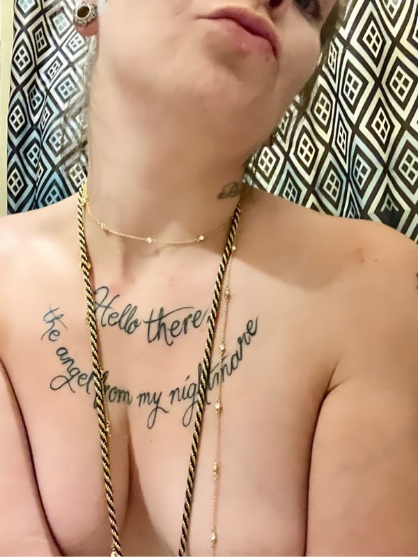 A woman with piercings and tattoos posing playfully in a bathroom setting, wearing layered necklaces.