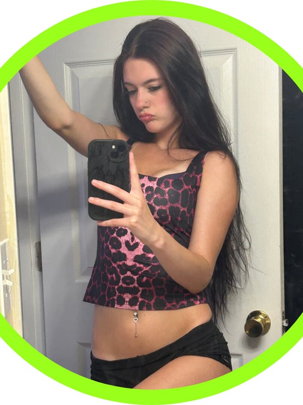A young woman with long dark hair taking a mirror selfie in a pink leopard print top and black shorts, standing in front of a door with a green circular frame.