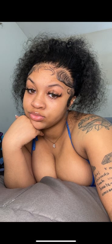 Young woman with curly hair and tattoos posing thoughtfully on a bed in a blue top.