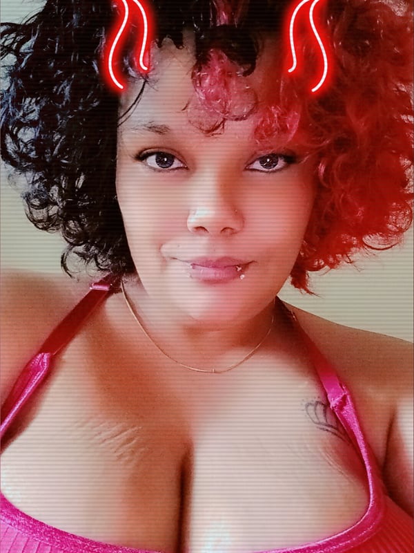 A woman with curly red and black hair and red devil horn effects smiles at the camera, wearing a pink top.
