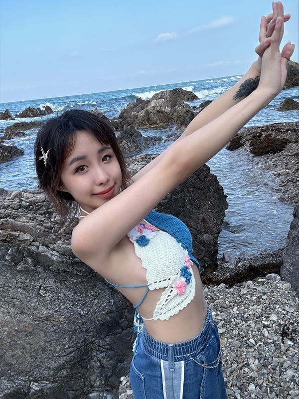 Young woman in a crochet top and denim shorts posing with arms raised on a rocky beach with ocean waves and blue sky.