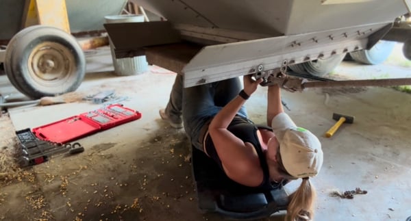 A woman in a black tank top and cap working on machinery in a workshop, holding a hammer and wearing work gloves.