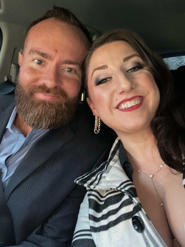 A bearded man in a suit and a woman with dark hair and earrings smile closely together inside a car.
