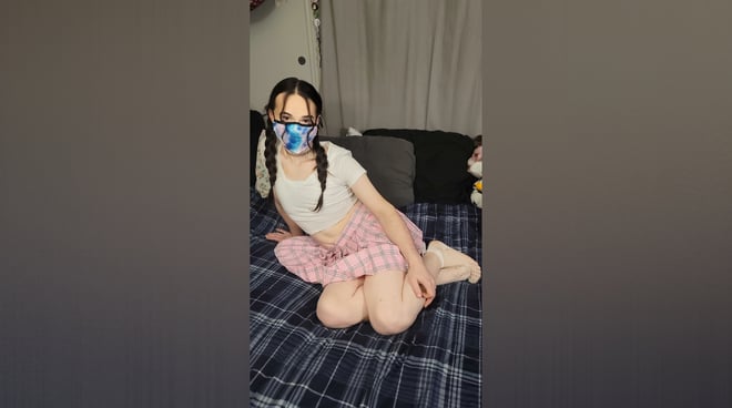 A person sitting on a bed in casual clothing with pigtails and a face mask, surrounded by plush toys and a curtained background.