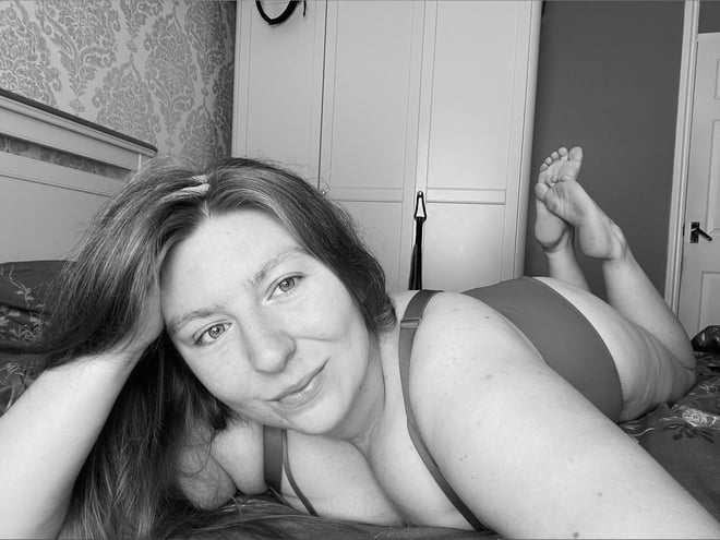 Woman with long hair lounging on a bed in a bedroom, smiling at the camera while wearing a swimsuit-style outfit.