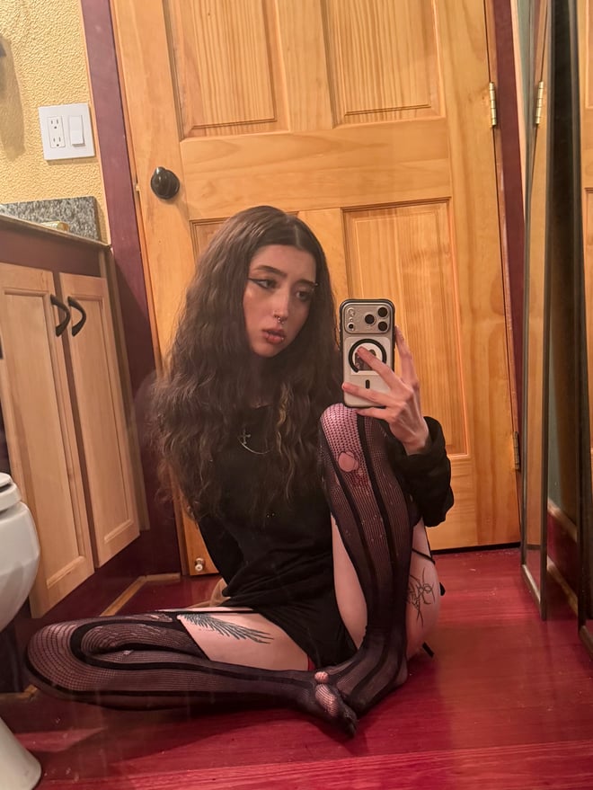 A young woman with long dark hair posing indoors near a doorway, wearing a white jacket over a lace top.
