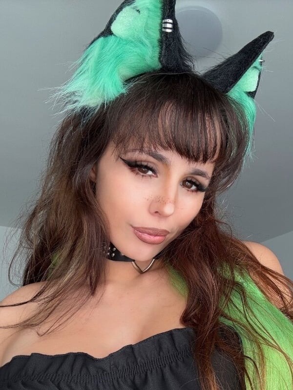 A woman with brown hair wearing green cat ears and a choker, smiling softly in a casual indoor setting.