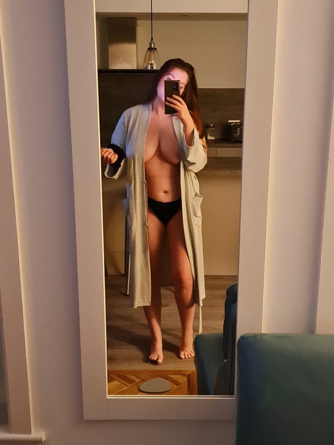 A woman taking a mirror selfie in a kitchen, wearing an open robe over underwear and smiling at the camera.