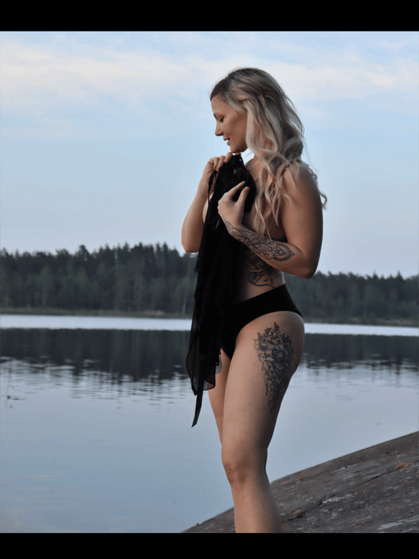 A blonde woman standing by the lake, holding a dark cloth over her chest while wearing swimsuit bottoms, with trees and water in the background.