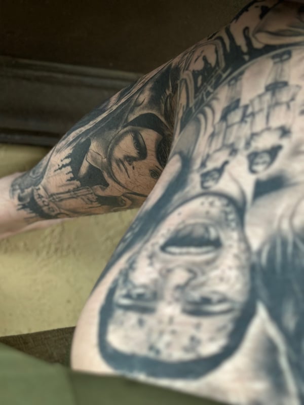 Close-up of intricate tattoos on a person's upper back and arm featuring skull and face designs in grayscale.