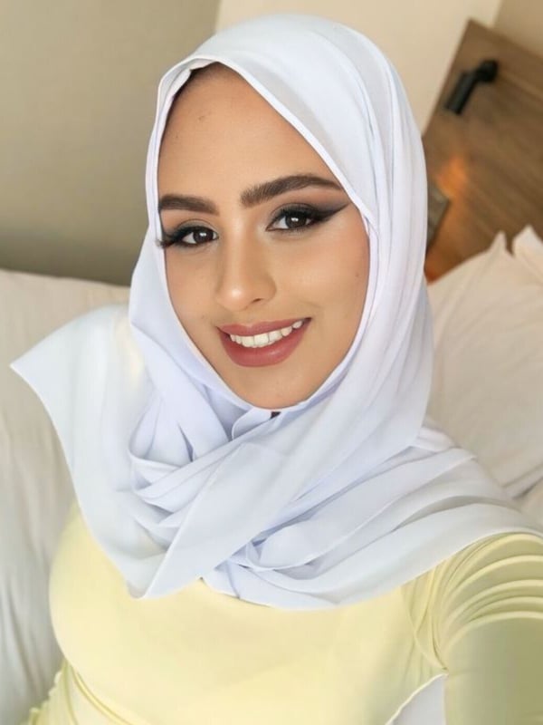 A smiling woman in a white hijab and yellow top takes a close-up selfie in a bedroom setting.