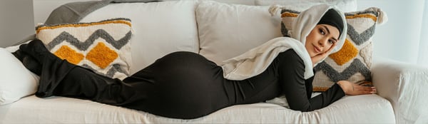 A woman in a white hijab and black dress reclining on a white couch with colorful patterned pillows in a bright room.