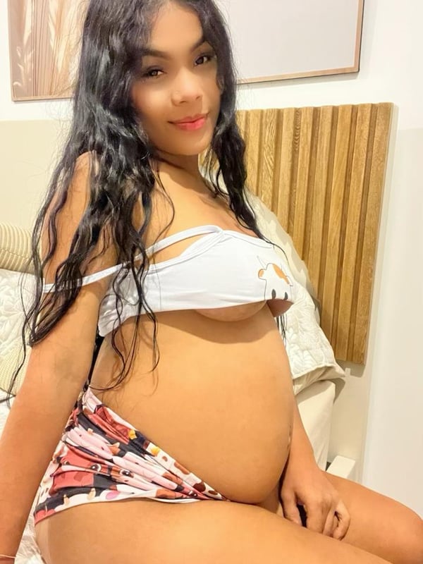 A pregnant woman taking a mirror selfie in a black bodysuit, standing in a modern bedroom.