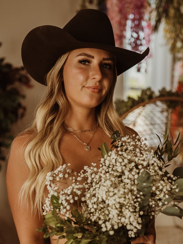 A smiling woman with long blonde hair wearing a cowboy hat, holding a bouquet of white flowers and greenery in a room with plants.