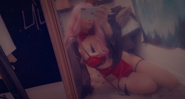A person with pink hair posing in red lingerie in front of a mirror in a bedroom setting.