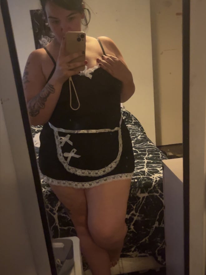 A woman with dark hair in a bun wearing a black maid-style outfit, taking a mirror selfie in a bedroom.