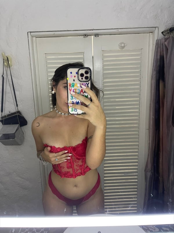 Young woman taking a mirror selfie in a red lace lingerie set inside a room with a wooden door.