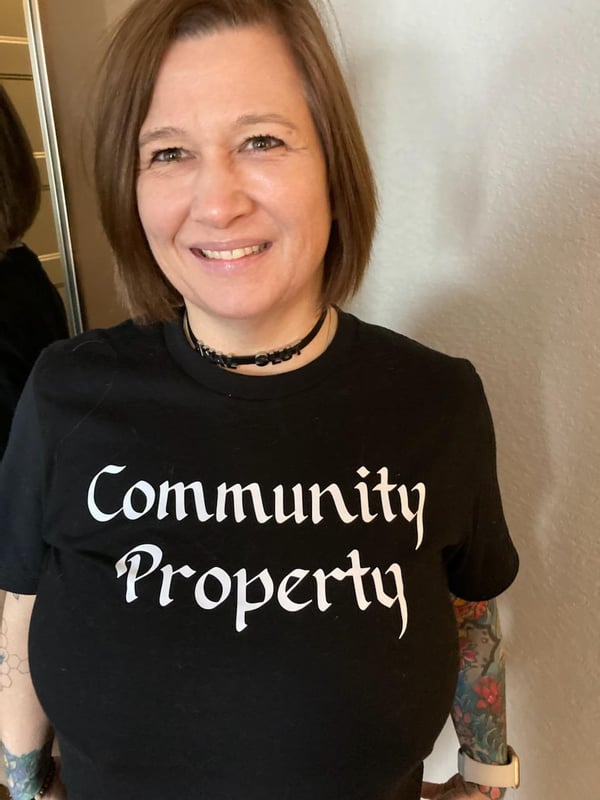 A smiling woman with brunette hair and tattoos wearing a black 'Community Property' t-shirt and choker necklace, standing confidently.