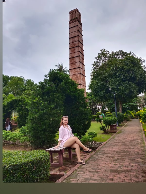 A woman in a light pink outfit sits on a bench in a lush garden with greenery and a tall brick chimney tower in the background under a cloudy sky.