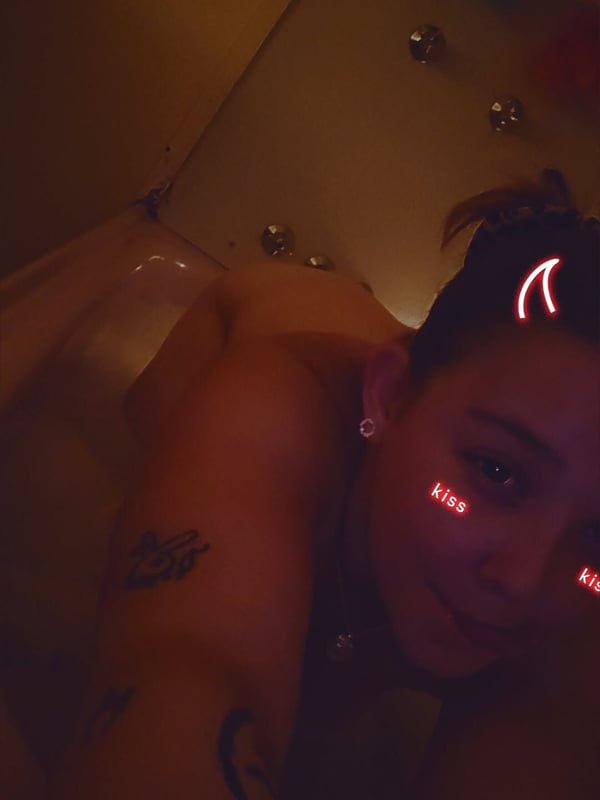 A woman with devil horns hair accessory and glowing red kiss marks on her cheeks, smiling playfully in a dimly lit room with tattoos on her shoulder.