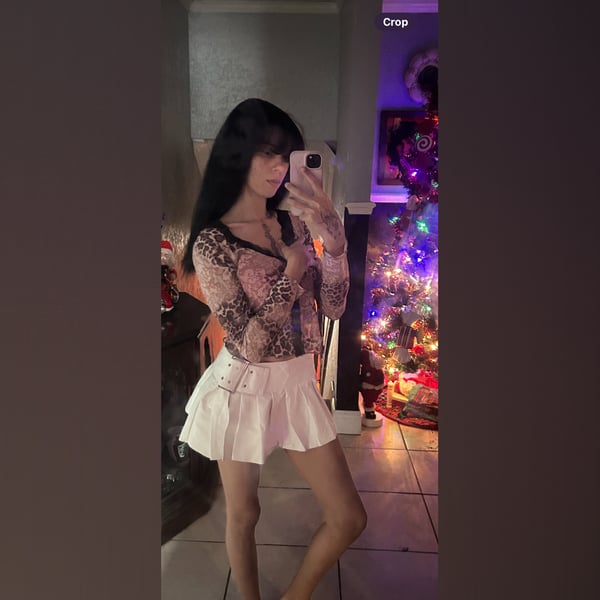 Young woman taking a mirror selfie in a sheer long-sleeve top and short white skirt, standing in front of a decorated Christmas tree in a living room.