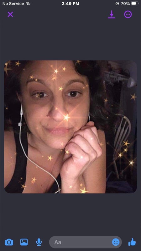 A young woman with curly dark hair wearing white earbuds and a black top, smiling with hand on cheek in a kitchen setting with sparkly filter effects.