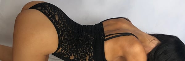 A woman posing on the floor in a black lace bodysuit against a plain background.