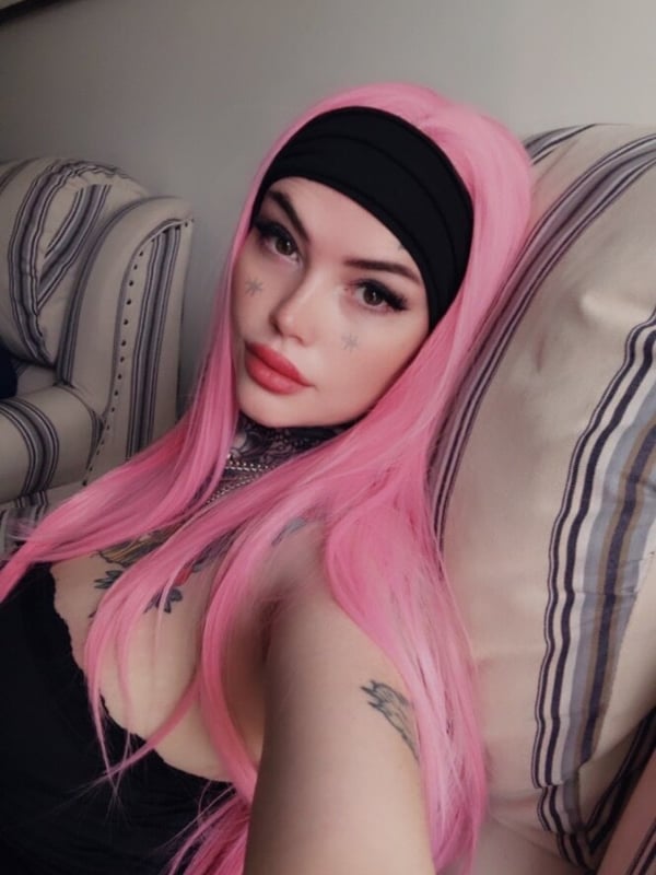 A woman with long pink hair and tattoos takes a selfie while seated on furniture wearing a black headband and top.