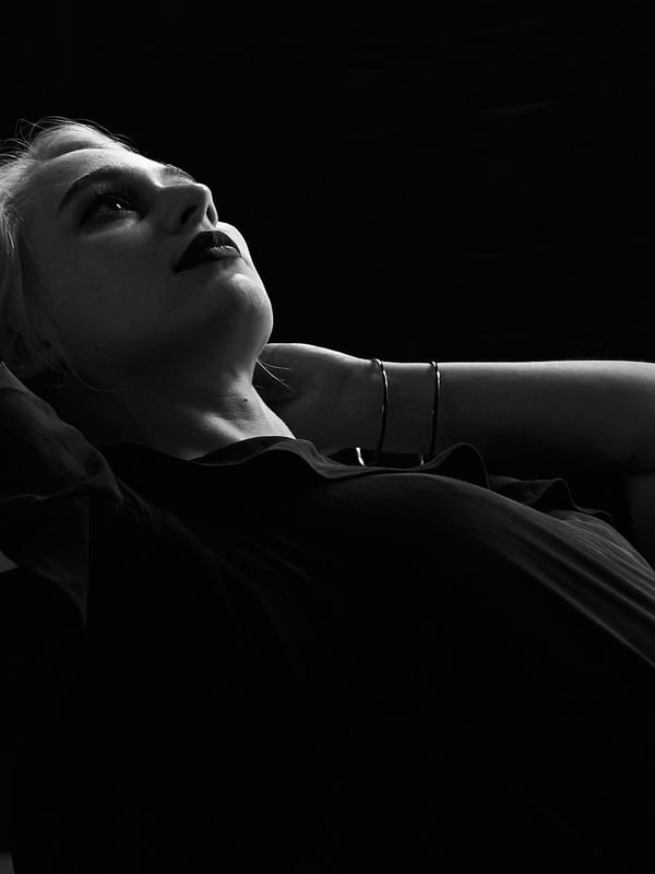A young woman with blonde hair tilting her head back and pursing her lips, posing in a black top against a dark background.