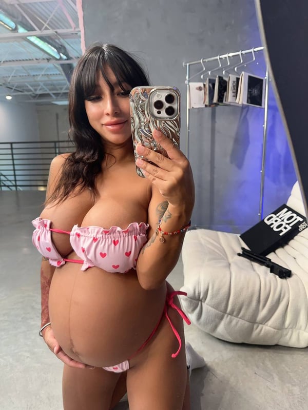 A pregnant woman standing outdoors in a leopard print bikini top, smiling with her hand resting on her belly near a window.