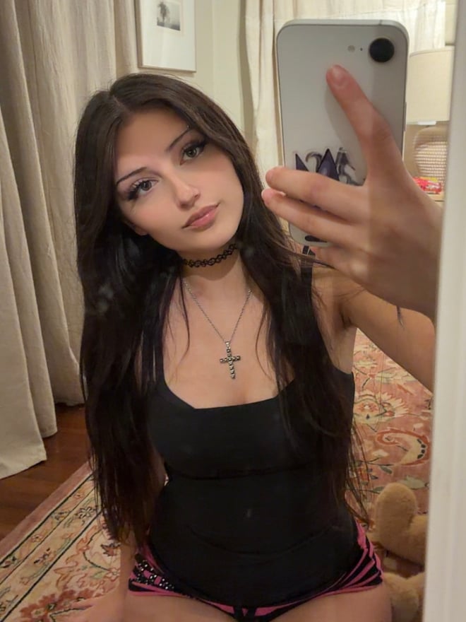High-angle selfie of a person with long dark hair, wearing a white lace-trimmed top and cross necklace, posing indoors on a patterned rug.