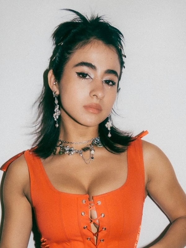 Young woman with dark hair styled messily, wearing an orange off-shoulder top with jewelry, posing confidently against a white background.