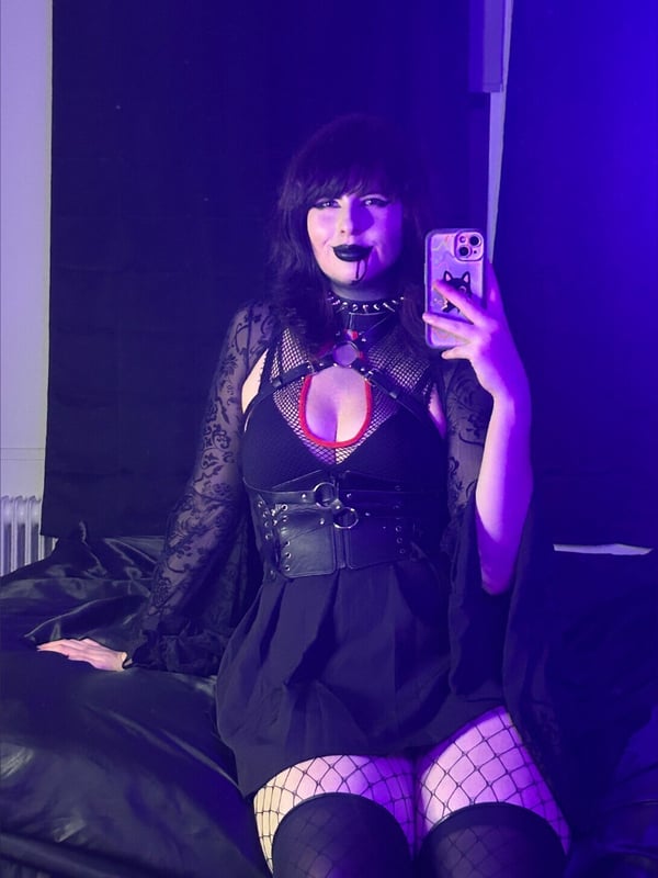 A woman with dark hair and bold makeup taking a selfie in gothic black lace clothing and fishnet stockings, illuminated by purple lighting.